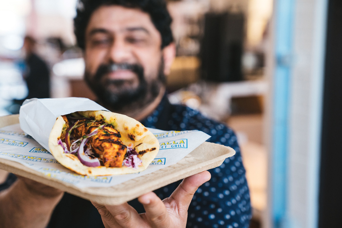 A man smiling in the background while he holds a hot buttered naan wrap