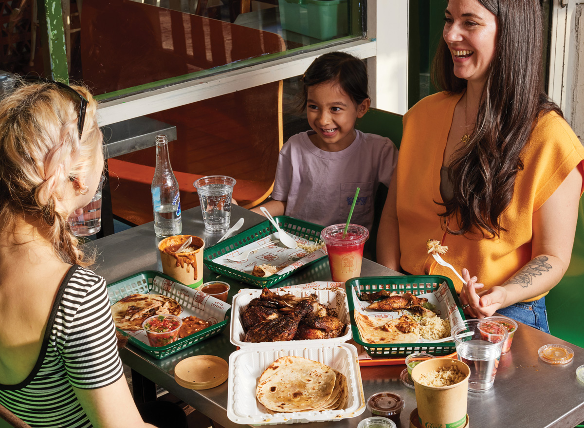 Friends laughing while sitting at a table with multiple trays of food and drinks