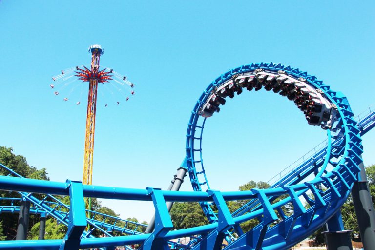 a dropping ride stands in the background while carts loop in a rollercoaster on the foreground rollercoaster