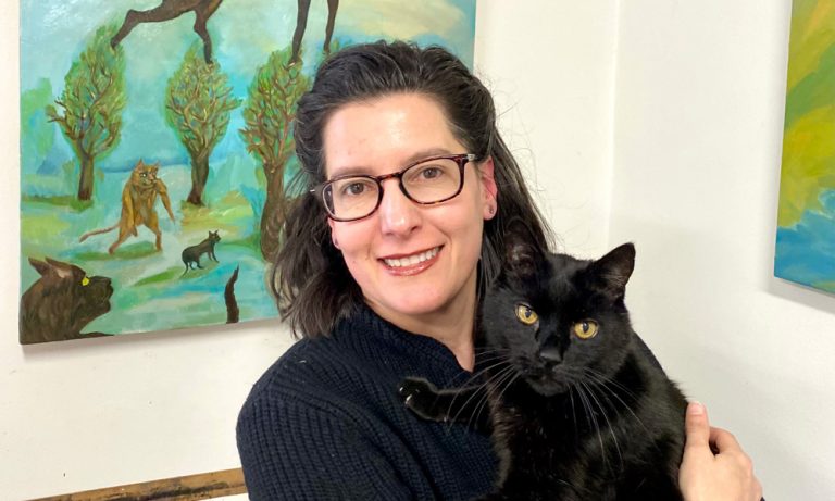 Artist Maria Korol smiles in front of her paints, holding a black cat in her arms