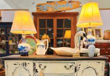 two antique lamps, a pitcher, books, and a bird decoration sit atop a distressed, white table