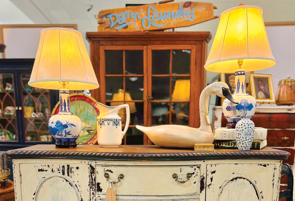 two antique lamps, a pitcher, books, and a bird decoration sit atop a distressed, white table