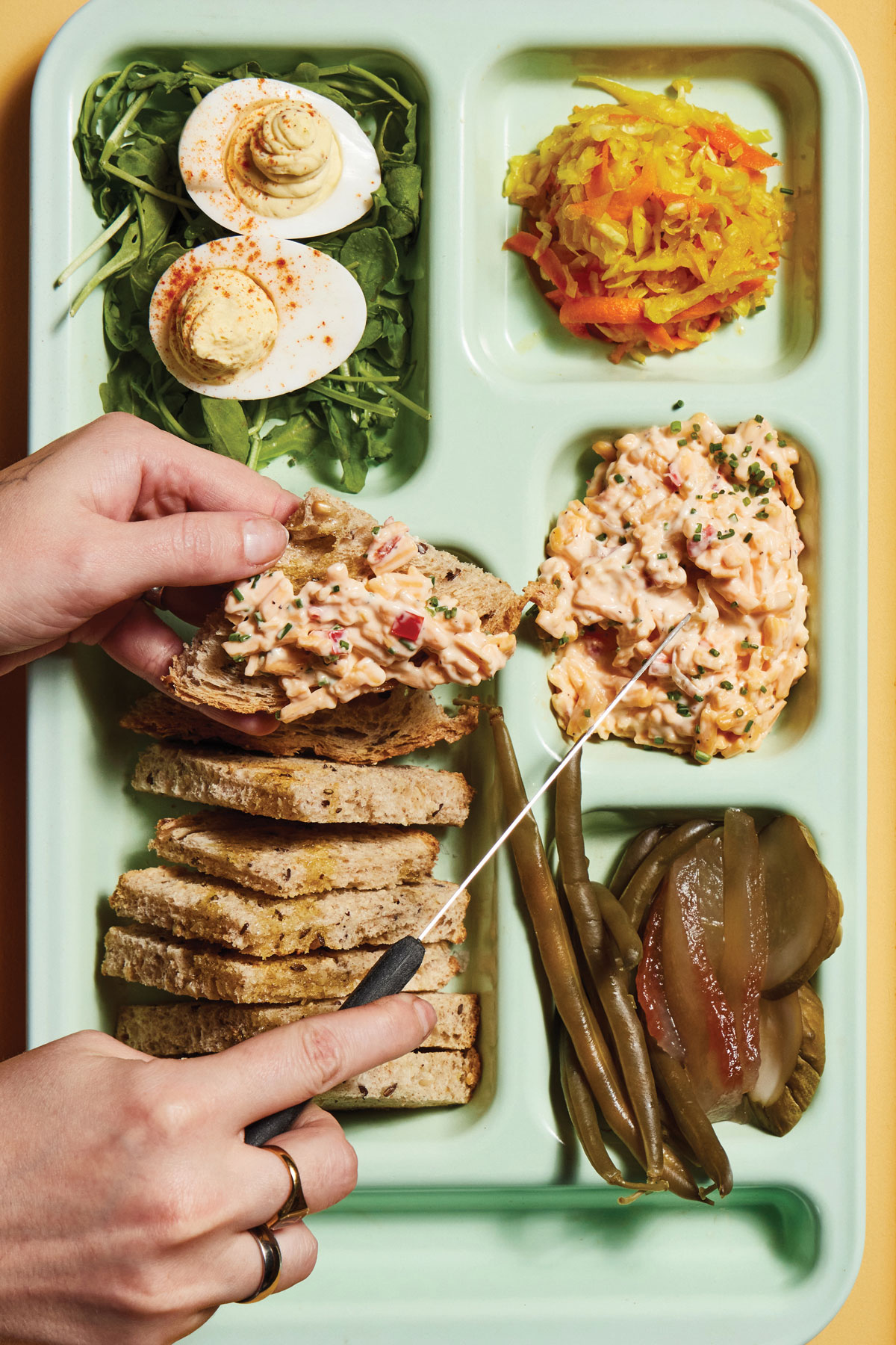 Two hands hover over a cafeteria-style tray of deviled eggs, chowchow, pickles, green beans, and more