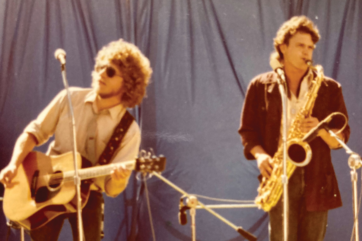 an older photo of Harrison playing the guitar on stage, next to a saxophone player