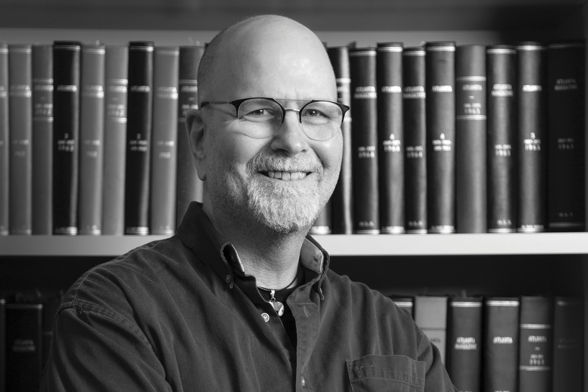 A black and white headshot of Scott Freeman crossing his arms and smiling in front of a bookshelf