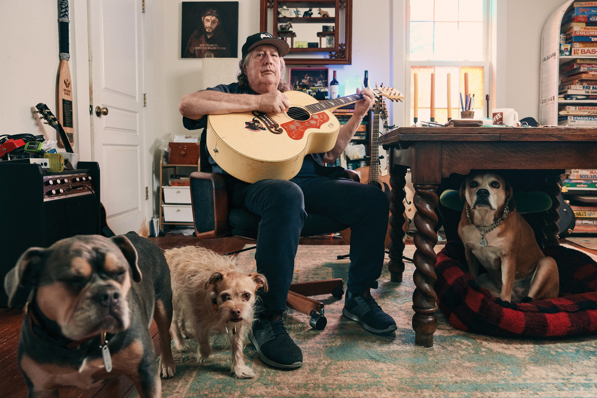 Kevn Kinney holds a guitar in his home, along with his three dogs surrounding him