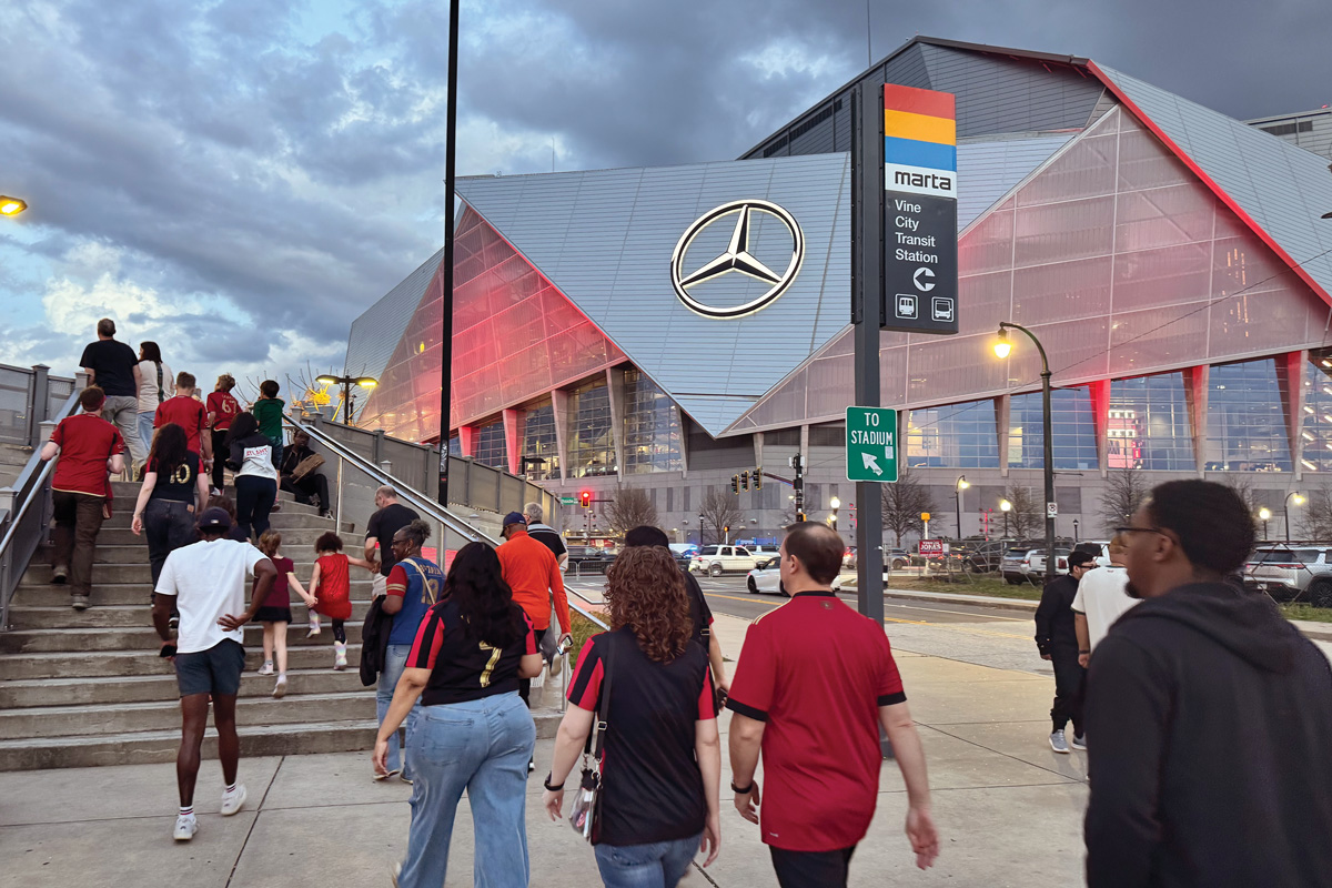 the outside of the Mercedes-Benz Stadium with people walking by a MARTA bus stop pole