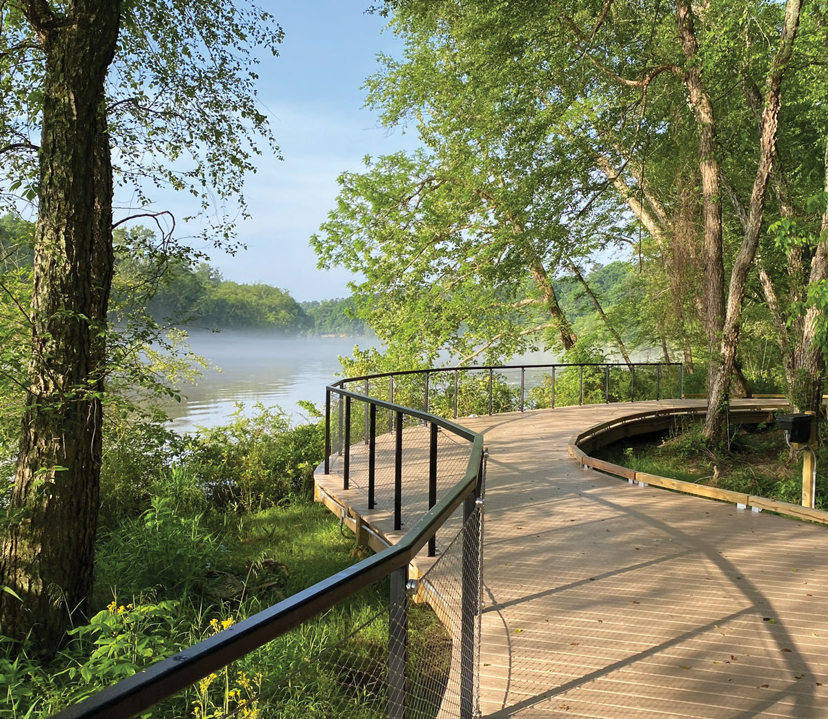 Trees and a lake surround a curved boardwalk on a blue sky day