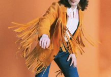 At Vintage by Judith, a clothing aficionado lives on through her 200,000-piece collection a person models blue jeans, a white button up, and a brown fringe suede jacket against a burnt orange backdrop