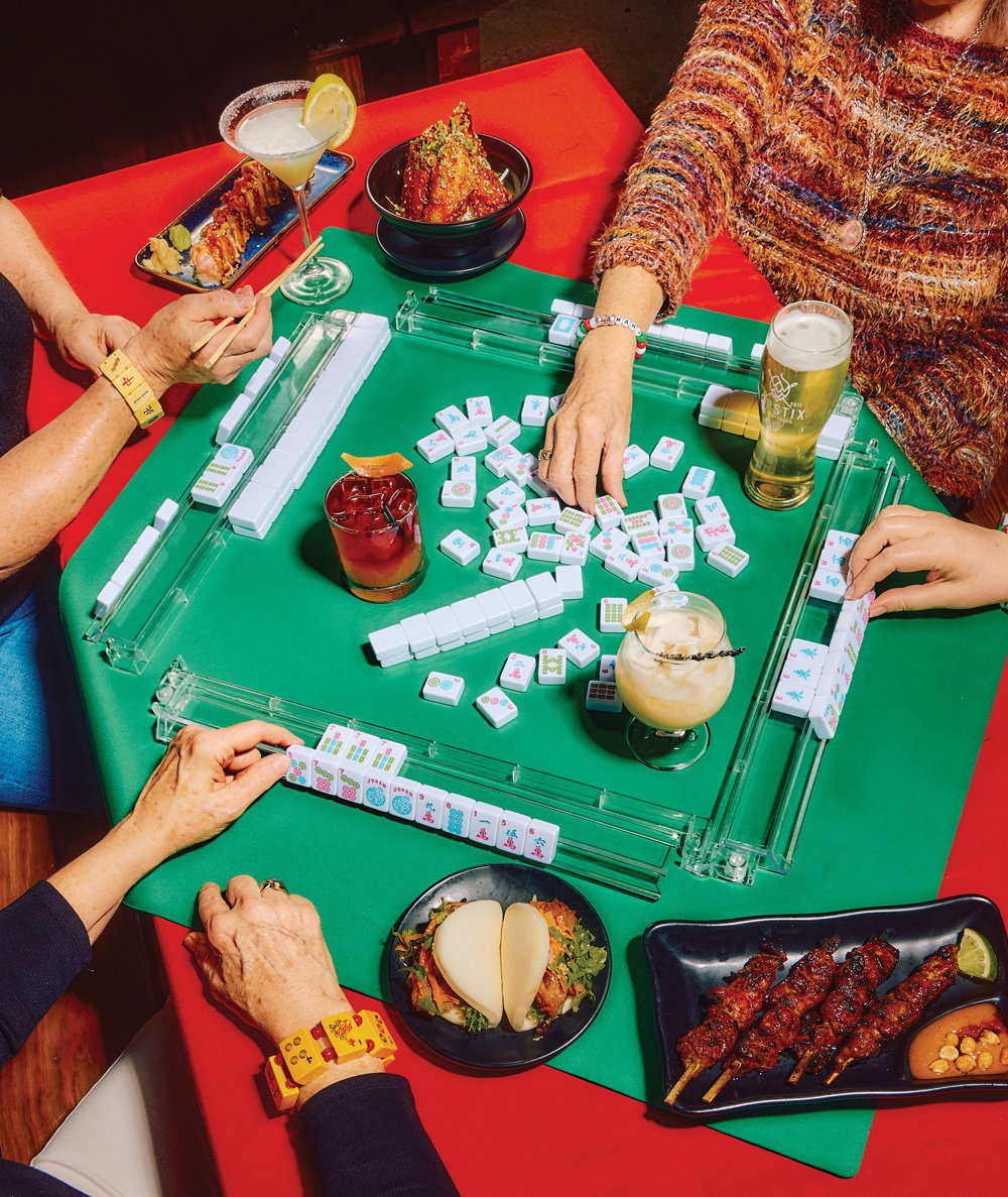 three women sit at a table playing Mahjong while enjoying food and drinks