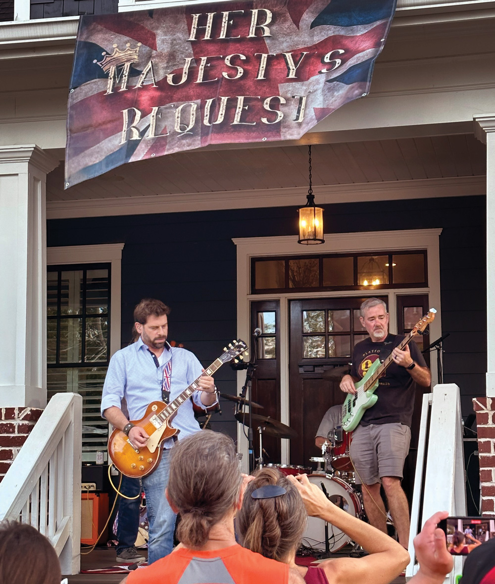 a band with a banner above them that reads "Her Royal Majesty" plays a show on a porch in front of an audience