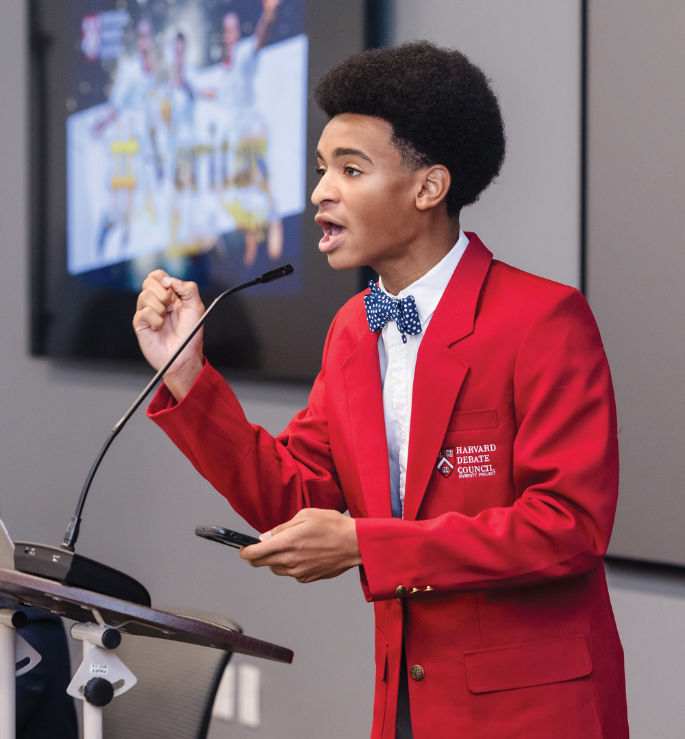 Isaiah Stephens at Harvard’s summer debate program, wearing a red blazer and speaking into a mic