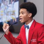 Isaiah Stephens at Harvard’s summer debate program, wearing a red blazer and speaking into a mic