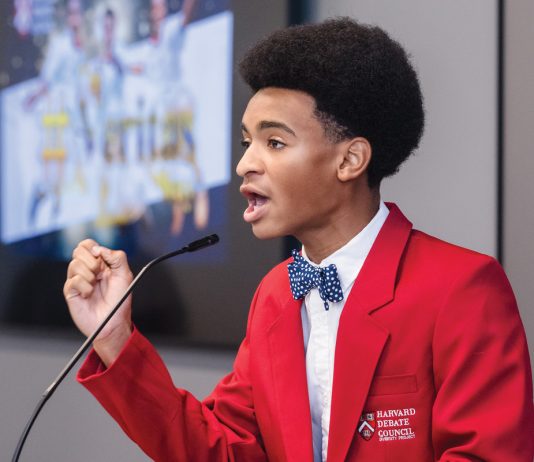 Isaiah Stephens at Harvard’s summer debate program, wearing a red blazer and speaking into a mic