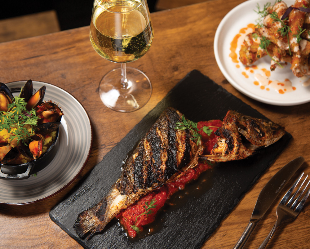 Grilled branzino with stewed tomatoes and mussels next to chicken wings over waffles