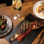 Grilled branzino with stewed tomatoes and mussels next to chicken wings over waffles