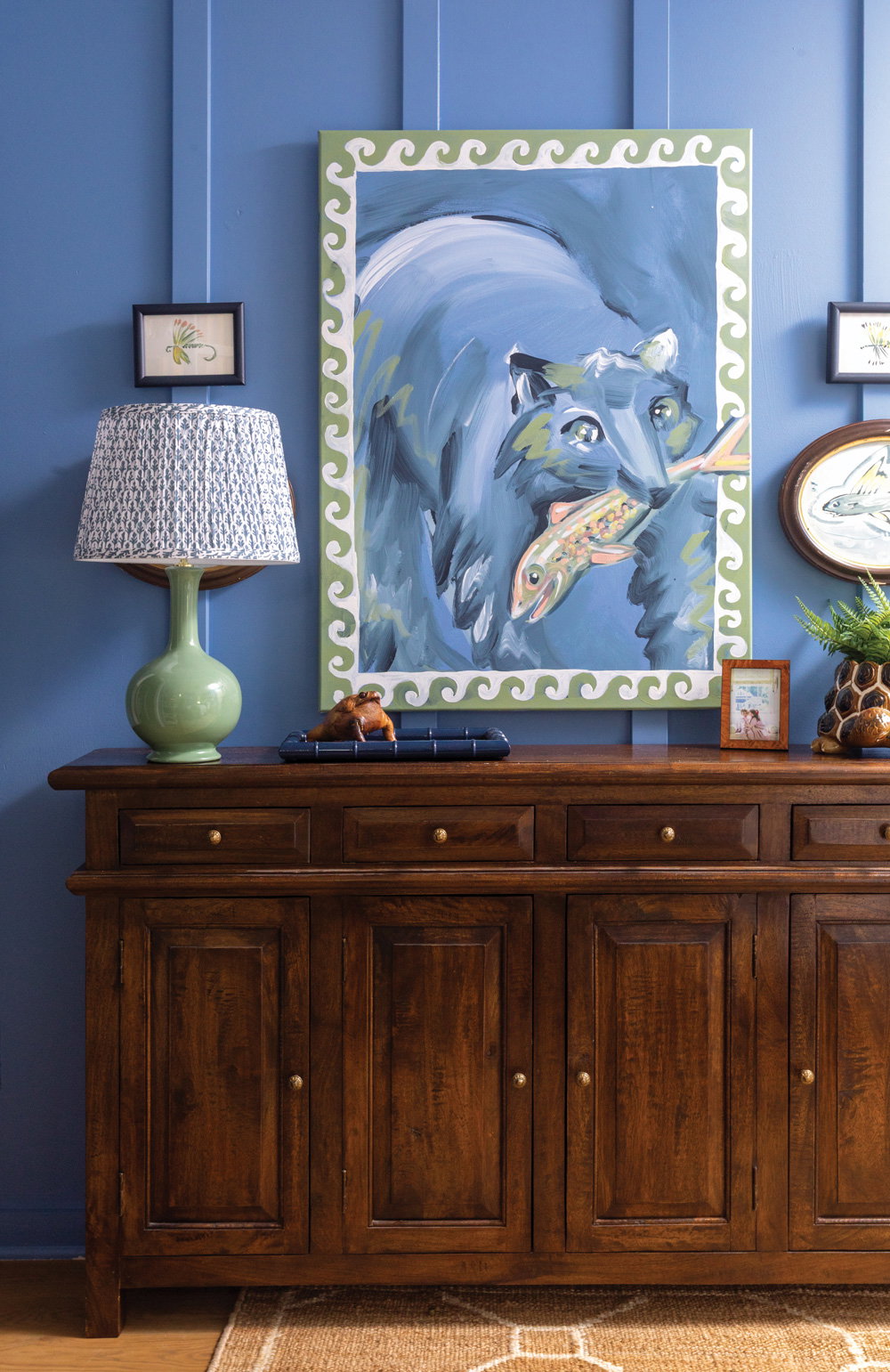 a deeper blue wall with an array of thrifted paintings over a wooden console table and green and beige lamp