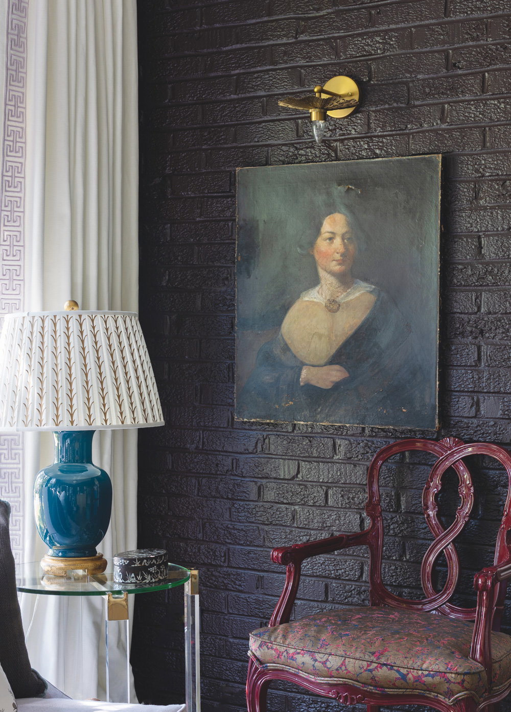deep chocolate brown walls with a hung portrait artwork on it, and a red leather chair and clear acrylic side table with a lamp atop