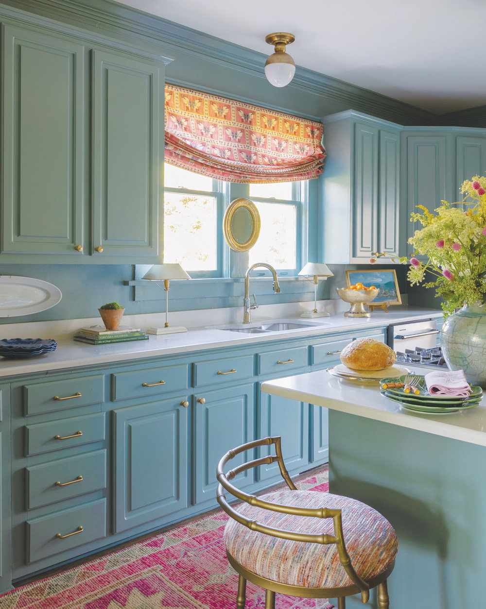 light blue cabinets with a patterned pinkish red window curtain and matching rug, and white marble countertops 