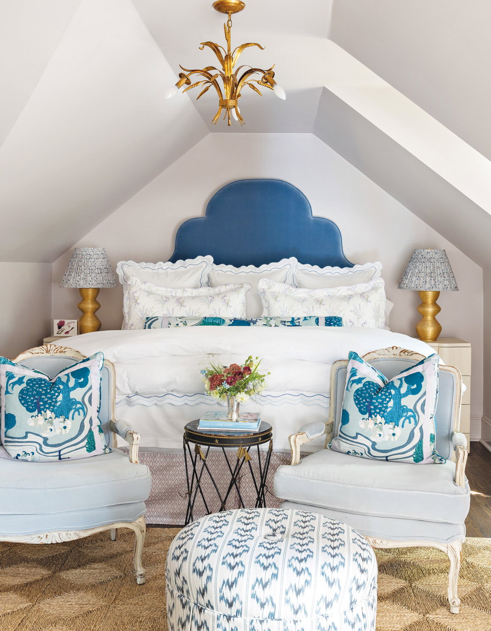 The bedroom's custom headboard wrapped in blue velvet. Brushed brass lamps and two light blue chairs with an ottoman stool at the foot of the bed