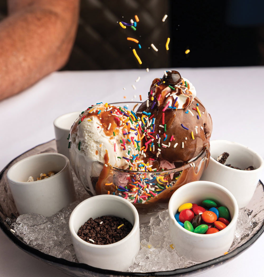 Sprinkles fall on a vanilla and chocolate ice cream sundae, surrounded by bowls of crushed Oreos, M&Ms, and other toppings