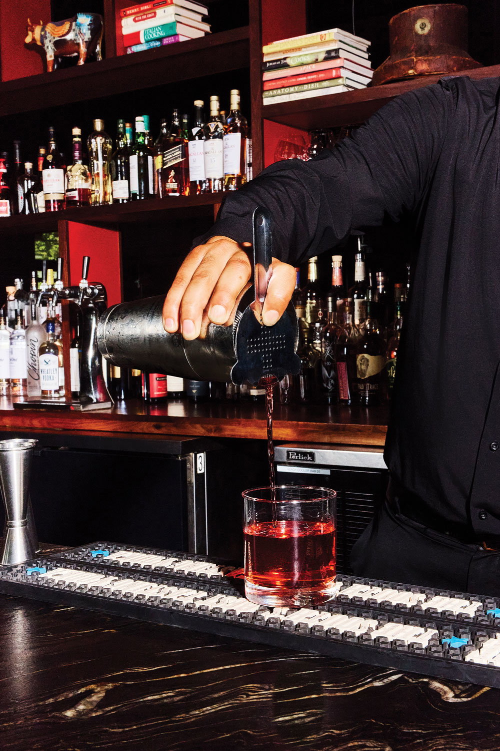 A bartender pours a cocktail with liquor bottles and trinkets lining the shelves behind him