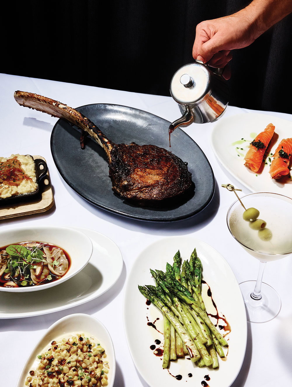 A server drizzles red wine jus on top of a tomahawk steak while a frosty martini and an assortment of sides surround it