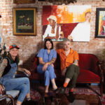 three people sit in mismatch couches against a brick wall with an array of paintings inside Last Dance