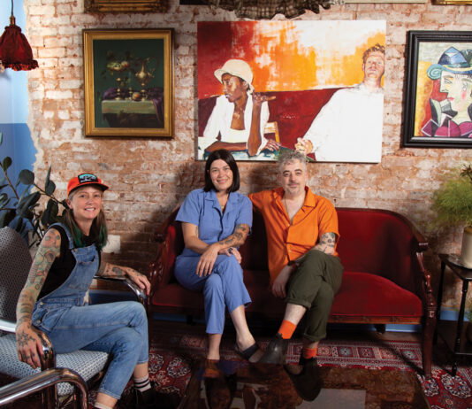 three people sit in mismatch couches against a brick wall with an array of paintings inside Last Dance