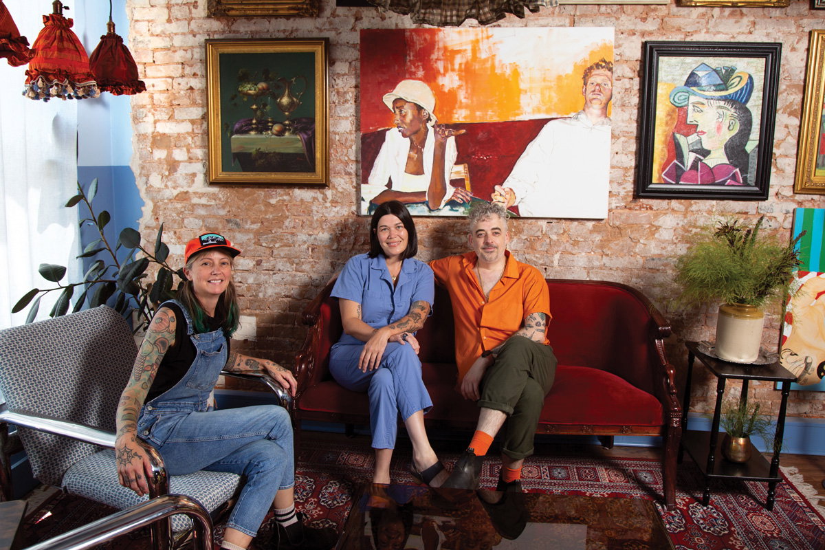 three people sit in mismatch couches against a brick wall with an array of paintings inside Last Dance